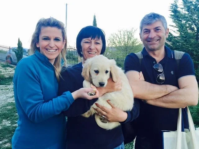 Tre persone sorridenti con un cucciolo di cane in un'area all'aperto con navi e alberi sullo sfondo.