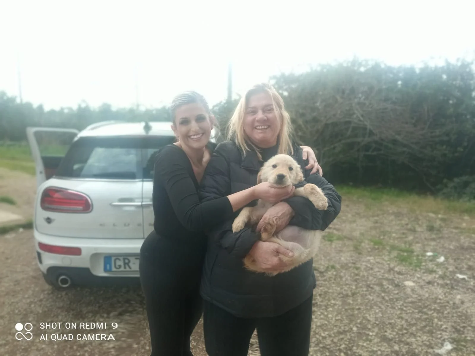 Due donne sorridenti sorridono con un cane di cucciolo in un'area all'aperto con un'auto bianca dietro e cespugli sullo sfondo.