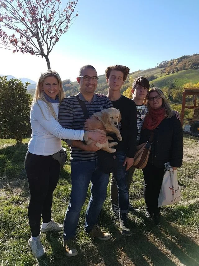 Gruppo di cinque persone che posano all'aperto con un cane dorato in un paesaggio collinare con alberi e montagne sullo sfondo, in una giornata soleggiata.