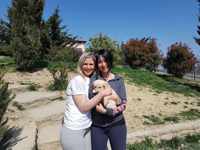 Due donne sorridenti che tengono un cucciolo di cane in un parco con alberi e un cielo azzurro.