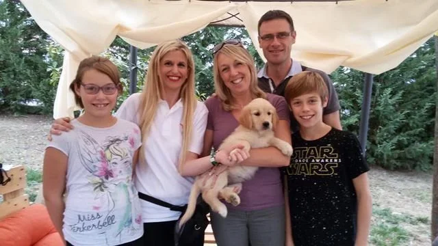Gruppo di cinque persone e un cucciolo di cane in una tendina all'aperto, con piante e natura sullo sfondo.