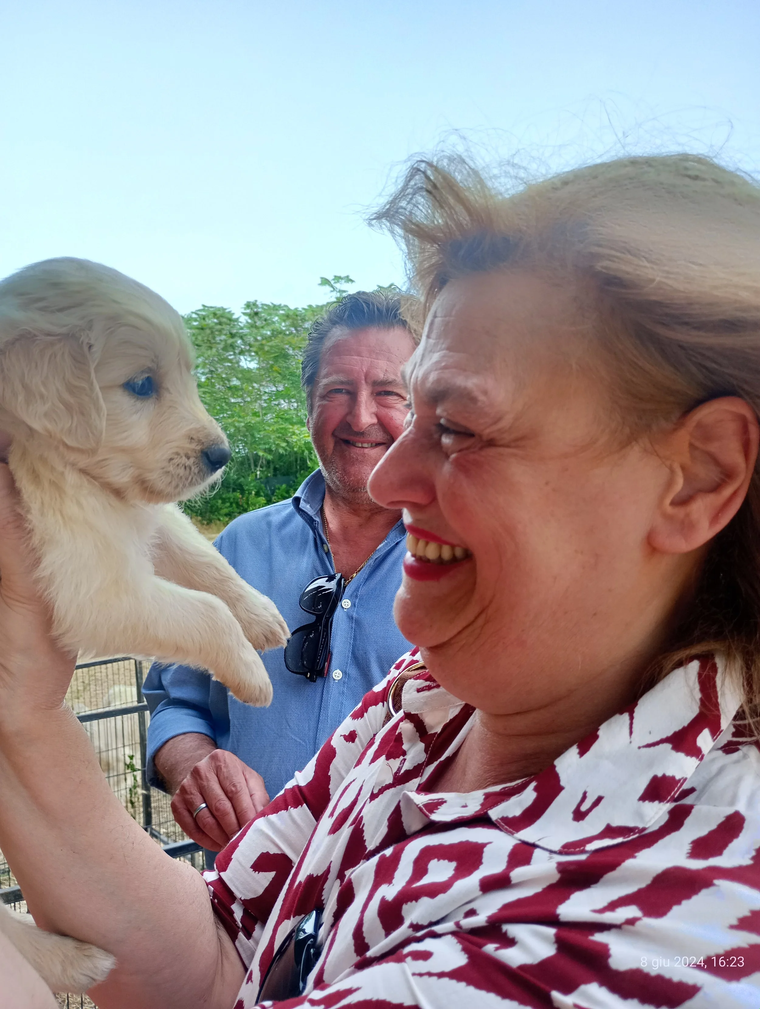 Un gruppo di persone all'aperto, si vede un cane cucciolo di colore chiaro che viene tenuto da una donna e sta guardando un'altra donna che sorride. Sullo sfondo ci sono alberi e un cielo sereno.