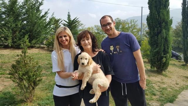 Tre persone sorridenti che tengono un cucciolo di cane in un giardino con alberi e colline sullo sfondo.