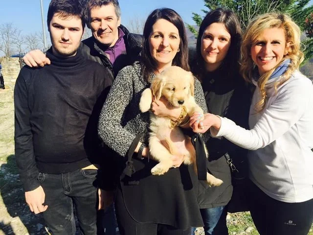Gruppo di cinque persone sorridenti con un cucciolo di cane dorato, in piedi all'aperto durante il giorno, con alberi e cielo azzurro sullo sfondo.
