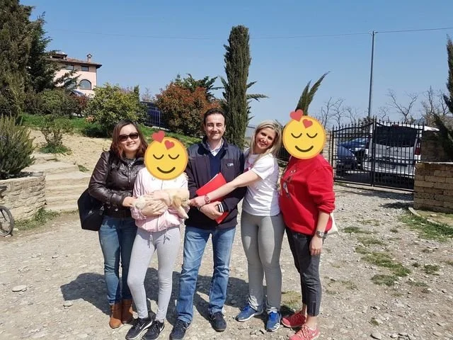 Gruppo di cinque persone che si abbracciano e sorridono all'aperto, con un cancello e alberi sullo sfondo.
