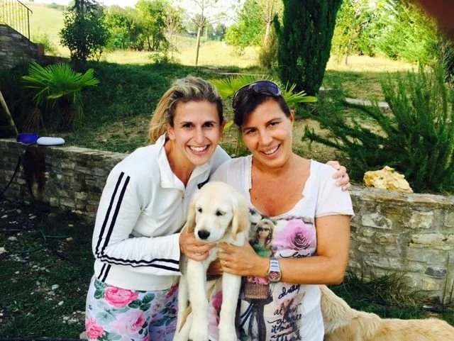 Due donne sorridenti che tengono un cucciolo di cane bianco in un giardino verde con piante e alberi