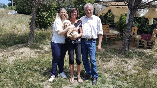 Tre persone sorridenti con un cane caldo in un giardino con alberi, erba e un gazebo