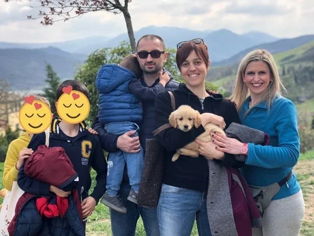 Una famiglia con due bambini e due donne, una delle quali tiene in braccio un cagnolino, in un paesaggio naturale con montagne sullo sfondo.