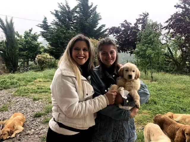 Due donne sorridenti che tengono un cagnolino tra loro, con altri cani sullo sfondo in un giardino verde con alberi e cespugli.