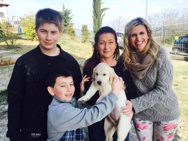 Una famiglia di quattro persone con un cane giallo, in un'area all'aperto con piante e un cancello in background.