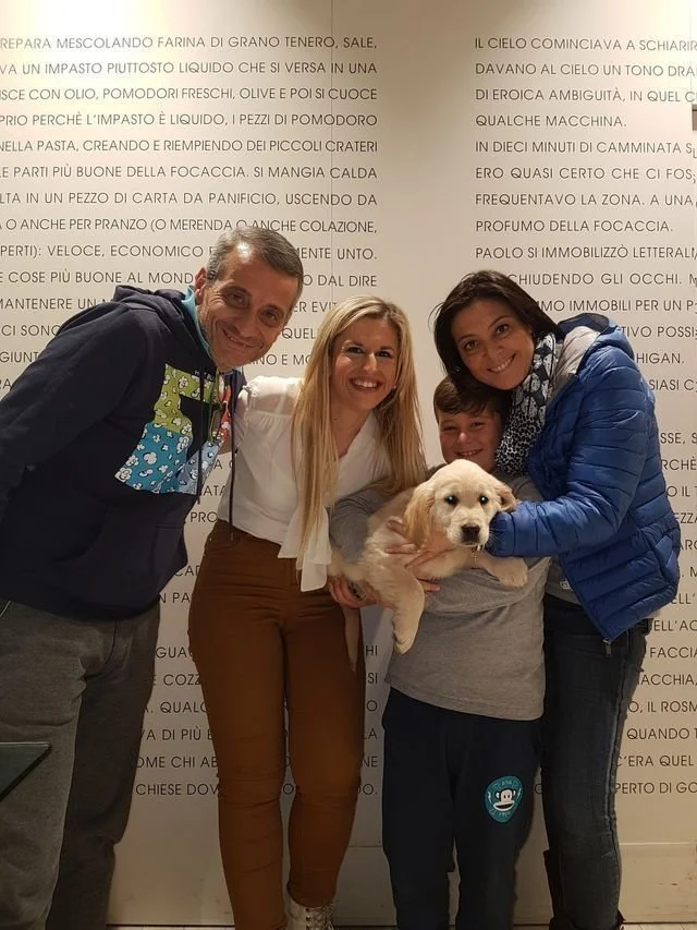 Quattro persone sorridenti che tengono un bambino e un cane bianco, davanti a un muro con testo in italiano