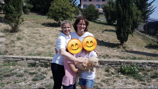 Tre donne sorridenti con un cane in un'area aperta con alberi e edera sullo sfondo.