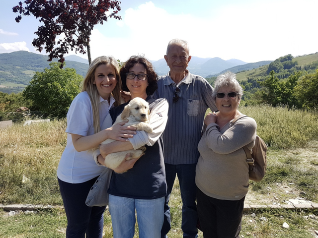 Quattro persone e un cane di fronte a un paesaggio collinare con alberi e montagne sullo sfondo in una giornata di sole.