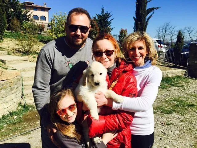 Un gruppo di quattro persone, tre donne e un uomo, sorridenti, tengono un cucciolo di cane bianco. Sono all'aperto, in un'area con piante e alberi, e il cielo è azzurro.