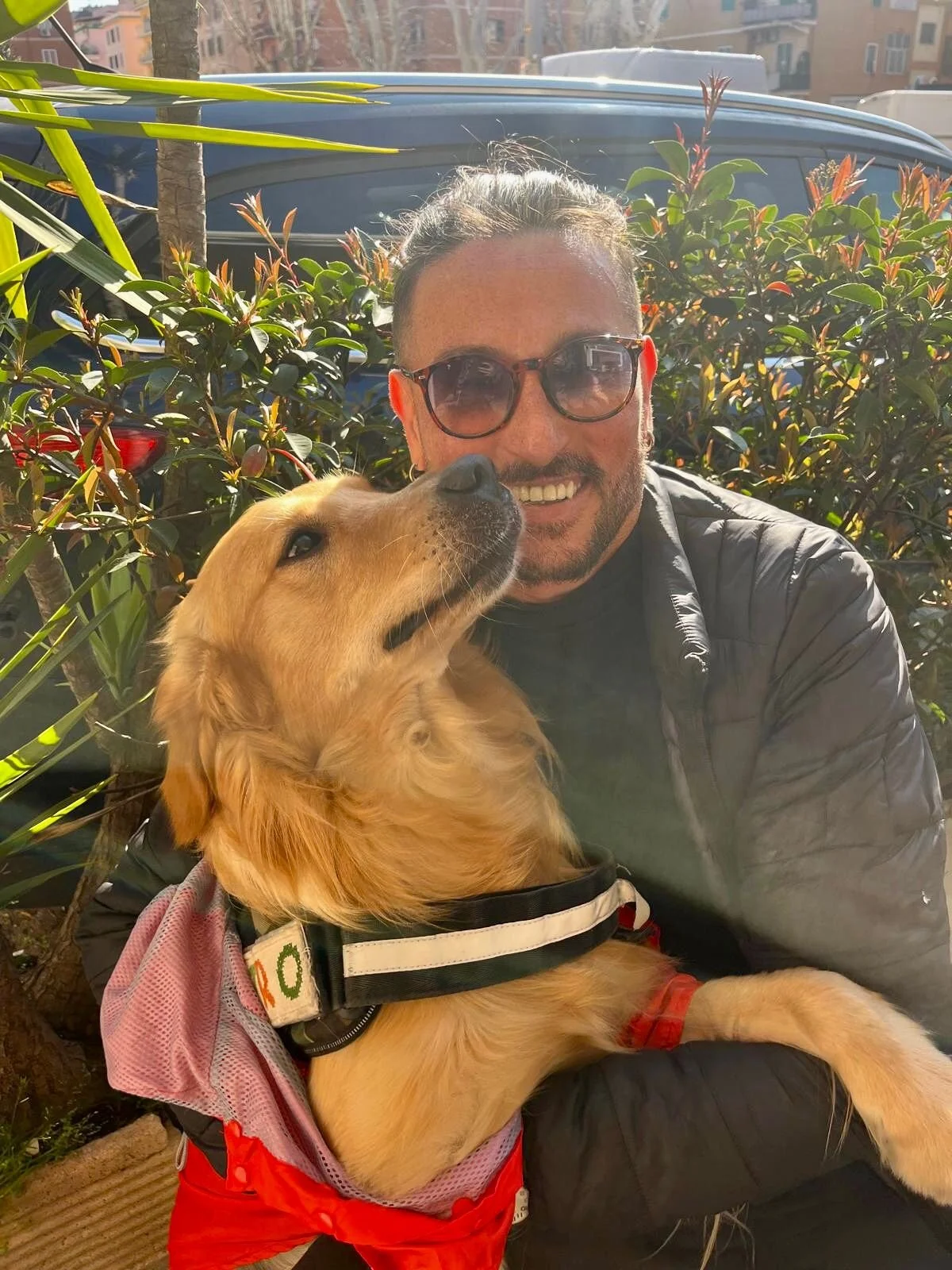 Un uomo con occhiali da sole e barba sorridente che abbraccia un cane golden retriever, seduti davanti a delle piante, in un ambiente all'aperto con edifici sullo sfondo.
