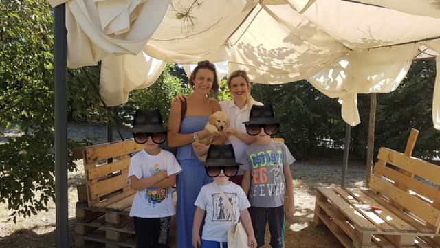 Tre donne adulte con tre bambini e un cucciolo di cane all'aperto sotto un tendone, con sedie di legno intorno.