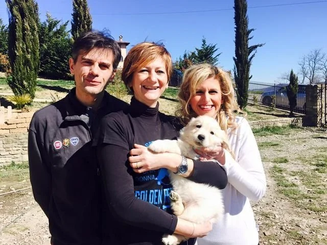 Tre persone sorridenti con un cane bianco in un cortile all'aperto con alberi e un cancello in background.