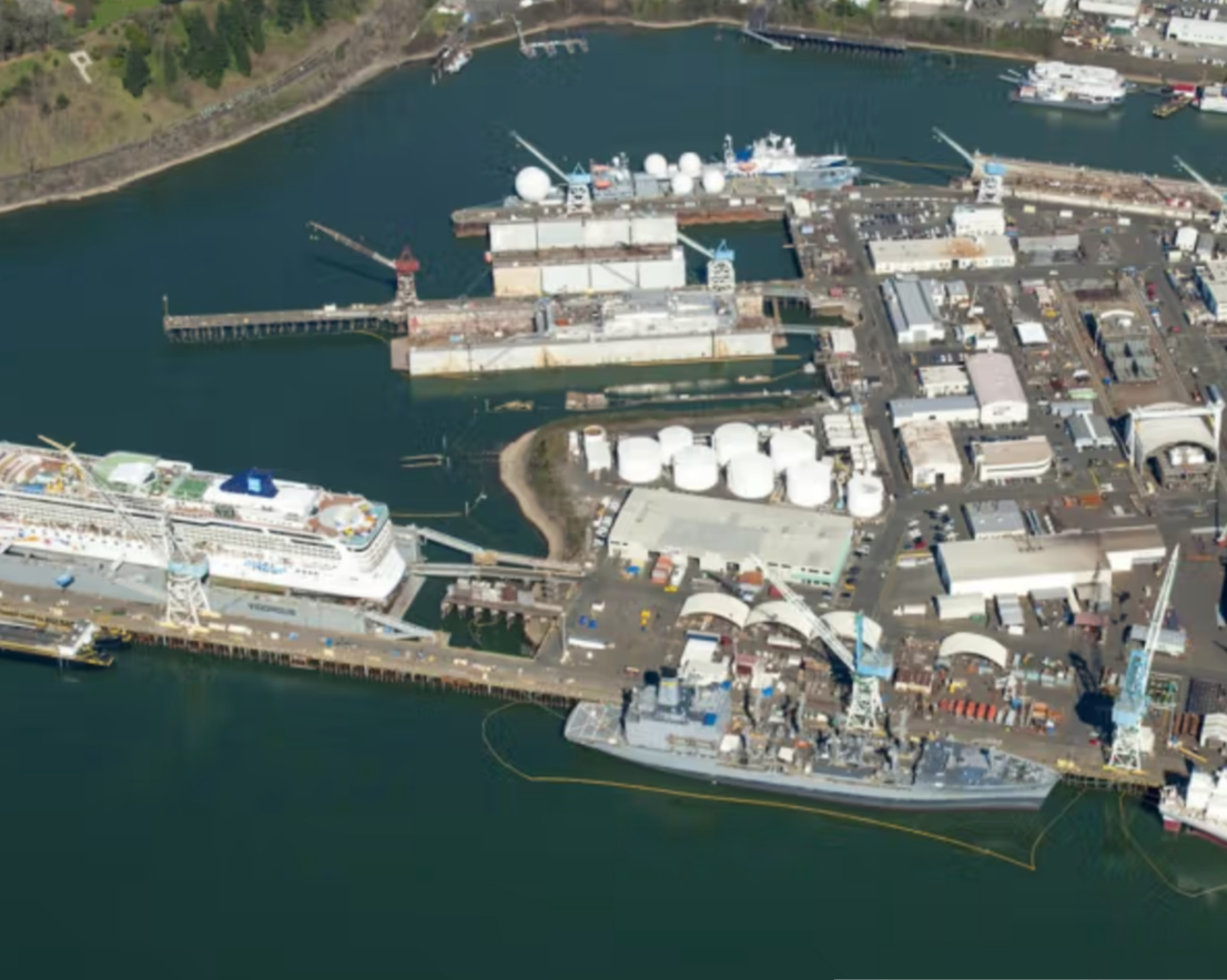 Aerial view of a busy harbor with a cruise ship docked, warehouses, storage tanks, and various industrial equipment.