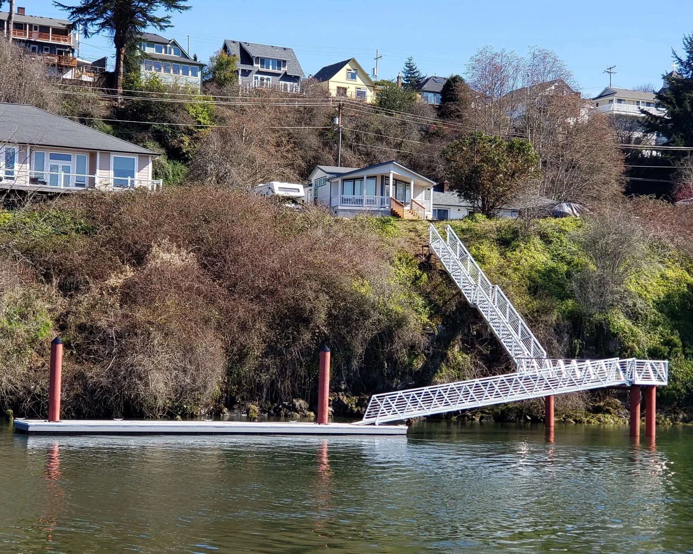 A white pedestrian bridge extending over a body of water with houses and trees on a hill in the background.