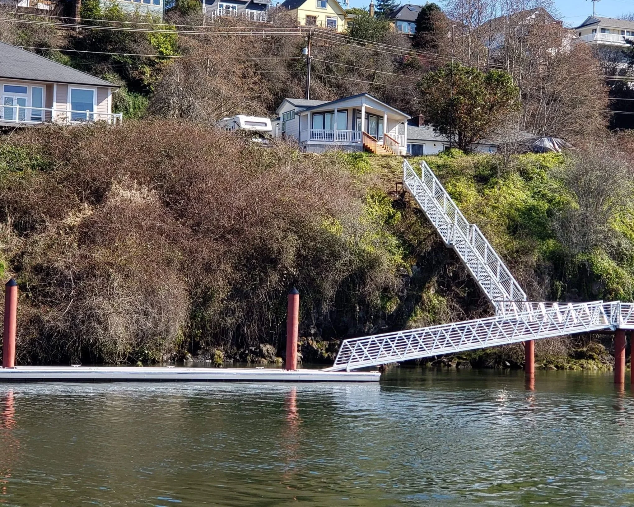 A white metal footbridge with stairs on the right side extends over a body of water, with red support poles and a railing. Multiple houses are on a hill in the background, overlooking the water, with trees and shrubs in the foreground.
