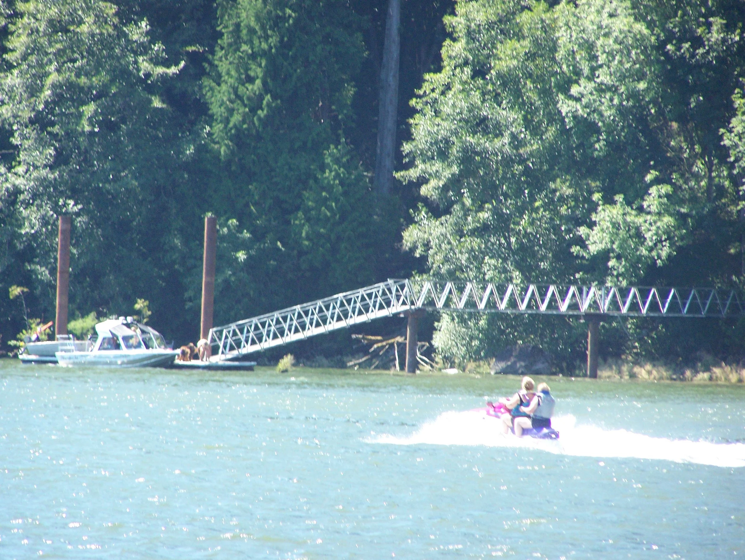 People enjoying a day on the water with boats, a dock, and a person riding a jet ski on a lake surrounded by trees.