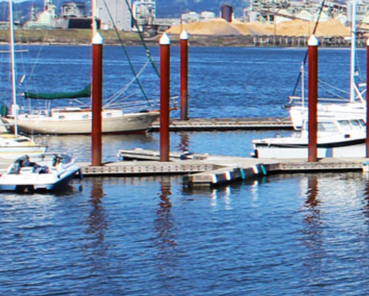 Boats docked at a marina with water and industrial buildings in the background.
