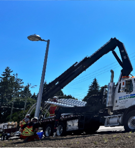 A large crane lifting a streetlamp during daytime with a clear blue sky and trees in the background.