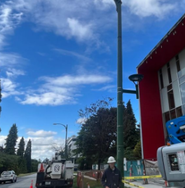 A construction worker wearing a yellow hard hat and dark clothing walking on a sidewalk beside a red building under a blue sky with clouds.