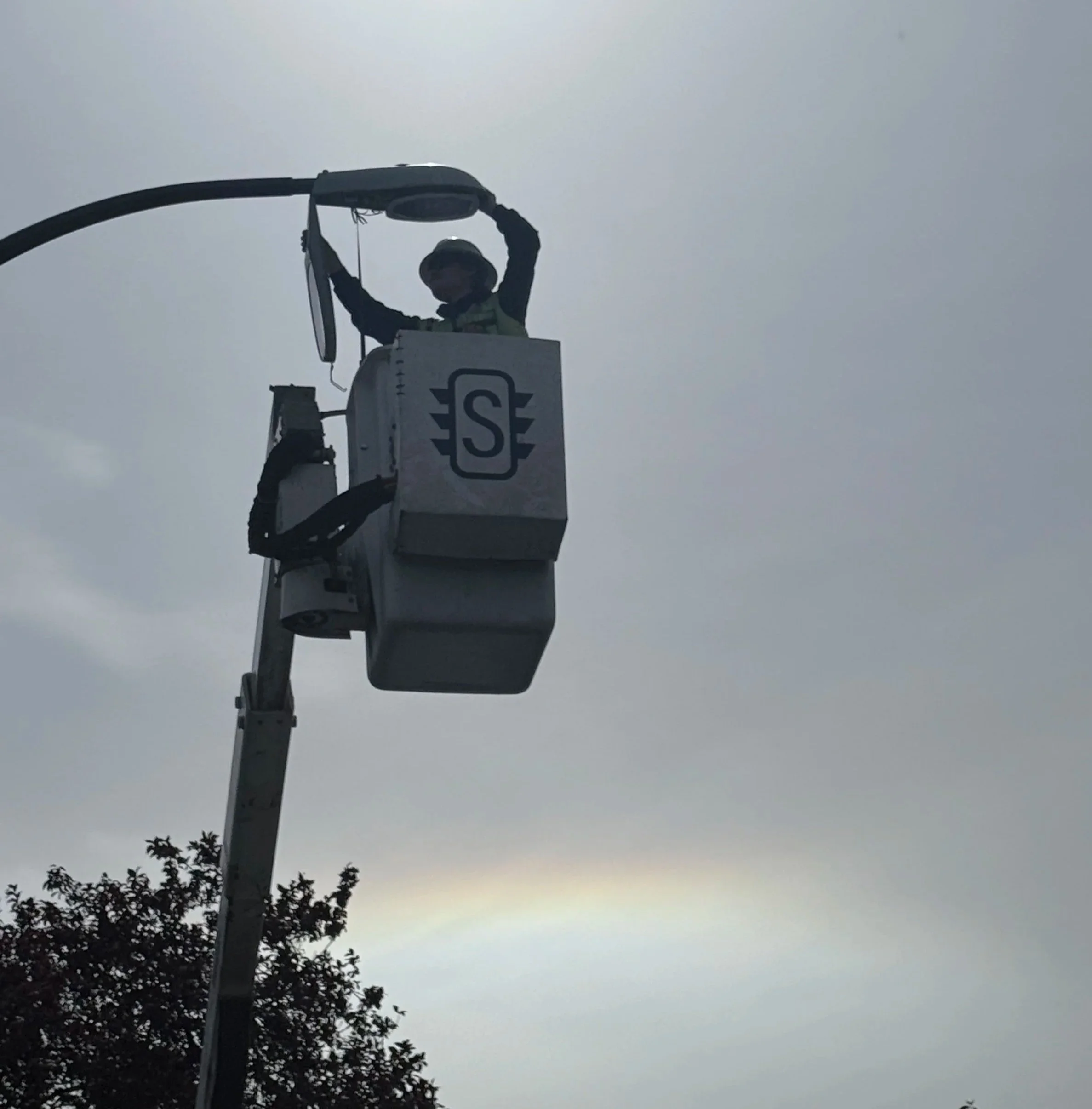 A worker in a bucket lift wearing safety gear, including a helmet, working on a streetlight against a cloudy sky with trees visible at the bottom of the image.