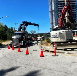 Construction site with a large black truck and an excavator, orange safety cones, and a tall building under construction in the background.