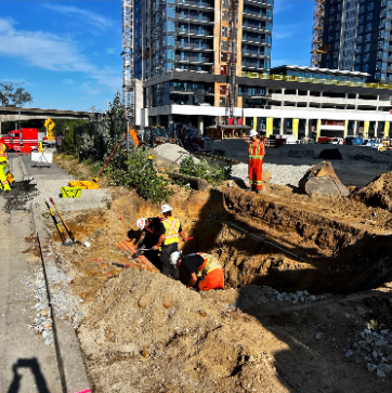 Construction workers digging a trench at a building site with tall modern buildings in the background.