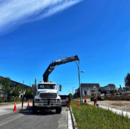 A utility truck with a crane lifting a streetlamp in a residential neighborhood with modern houses and a clear blue sky.