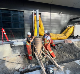 Two construction workers in safety gear working on a construction site with large yellow ventilation ducts and building materials.