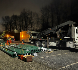 Construction workers setting up green metal framework in a nighttime outdoor area with trees in the background