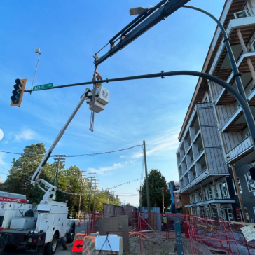 Construction site with a crane lifting a large light fixture, utility trucks, red safety fencing, and a multi-story building under construction against a blue sky.