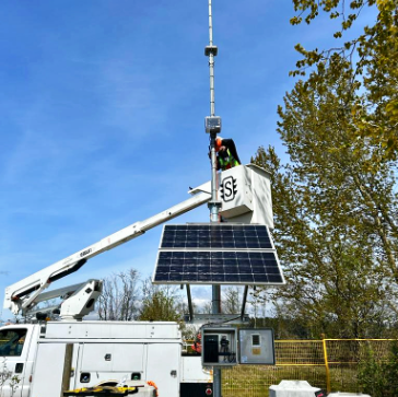 A worker installing a solar panel on a tall pole using a bucket truck in an outdoor setting with trees and a blue sky.