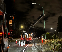 Nighttime scene of a street with a damaged streetlight leaning over the road. Emergency vehicles and cones are present, suggesting a recent accident or repair work.