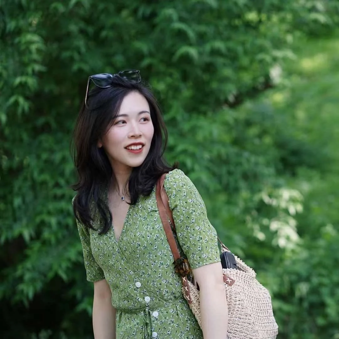 A young woman with black hair and sunglasses on her head smiling outdoors in front of green foliage, wearing a green floral dress and carrying a woven bag.