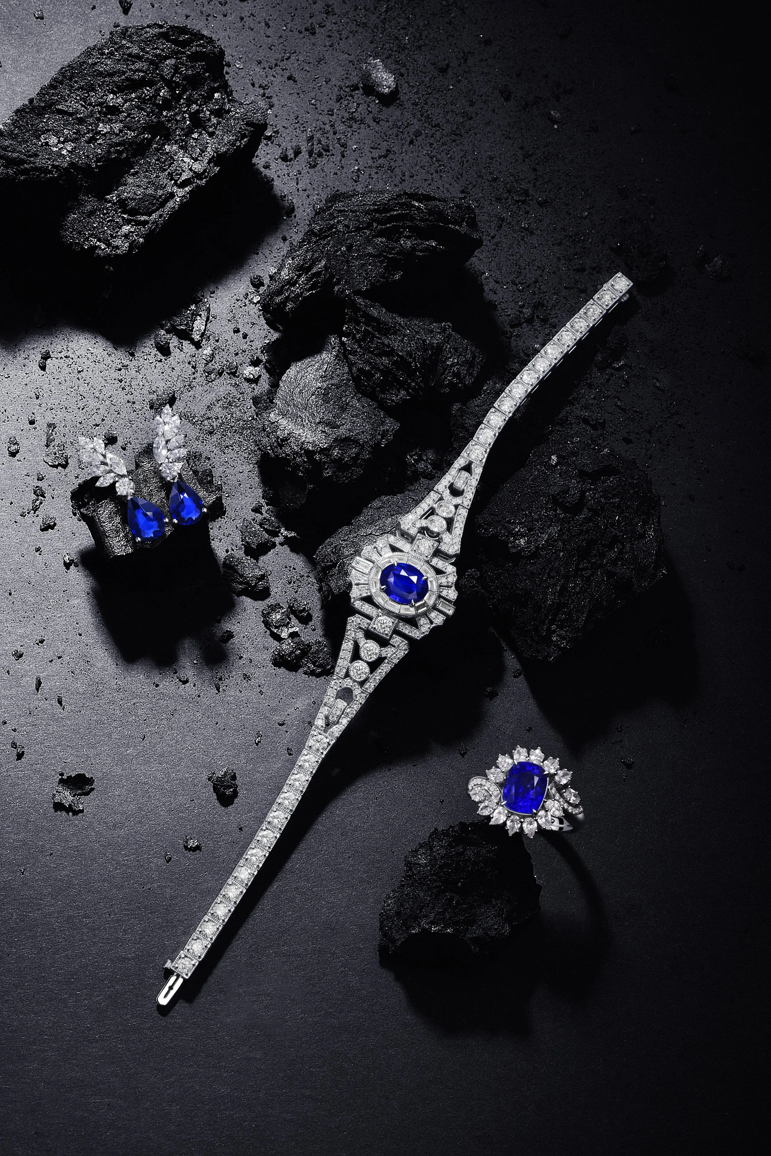 Jewelry set includes a diamond bracelet, earrings, and a ring, all featuring large blue gemstones and smaller diamonds, placed on a black textured surface with rocks around.