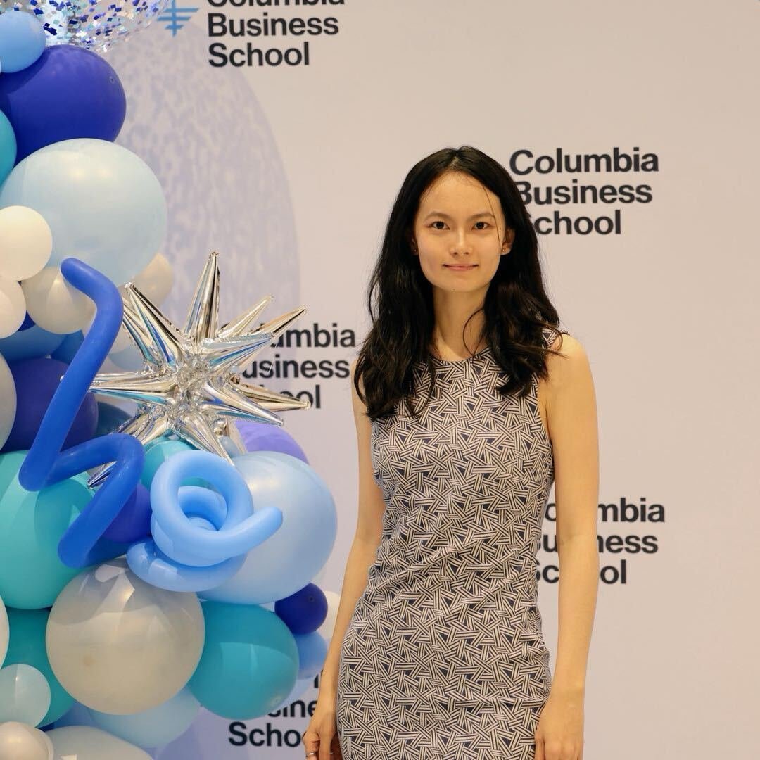 Young woman with long dark hair wearing a sleeveless patterned dress standing in front of a backdrop with 'Columbia Business School' written multiple times, next to a balloon arrangement with blue, white, and purple balloons and a silver star-shaped balloon.