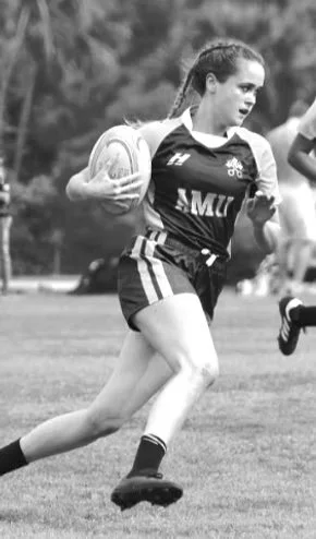 Female rugby player running with ball on the field in black and white.