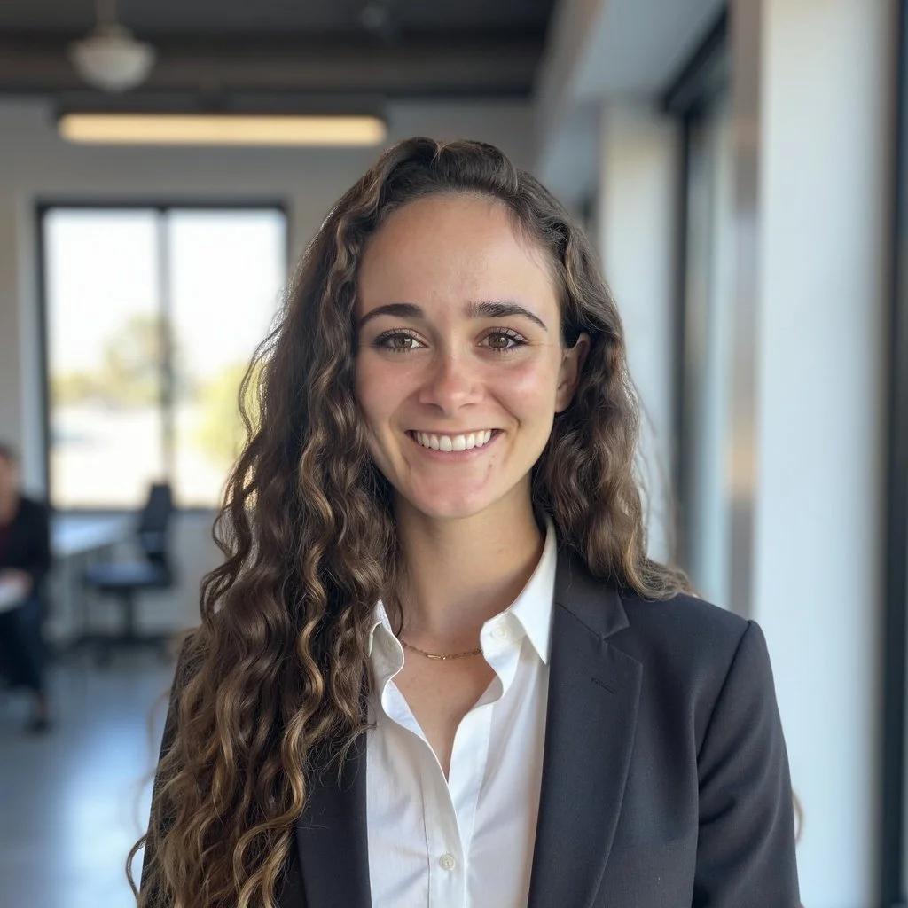A young woman with long, curly brown hair smiling in an office setting, dressed in a business suit with a white shirt.