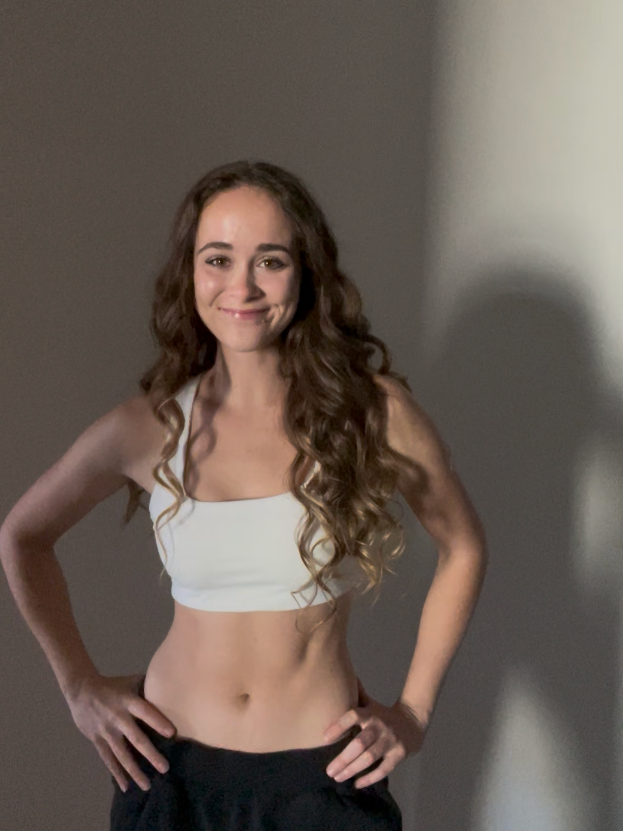 A young woman with long curly hair and a smile, standing against a plain wall, wearing a white crop top and black pants.