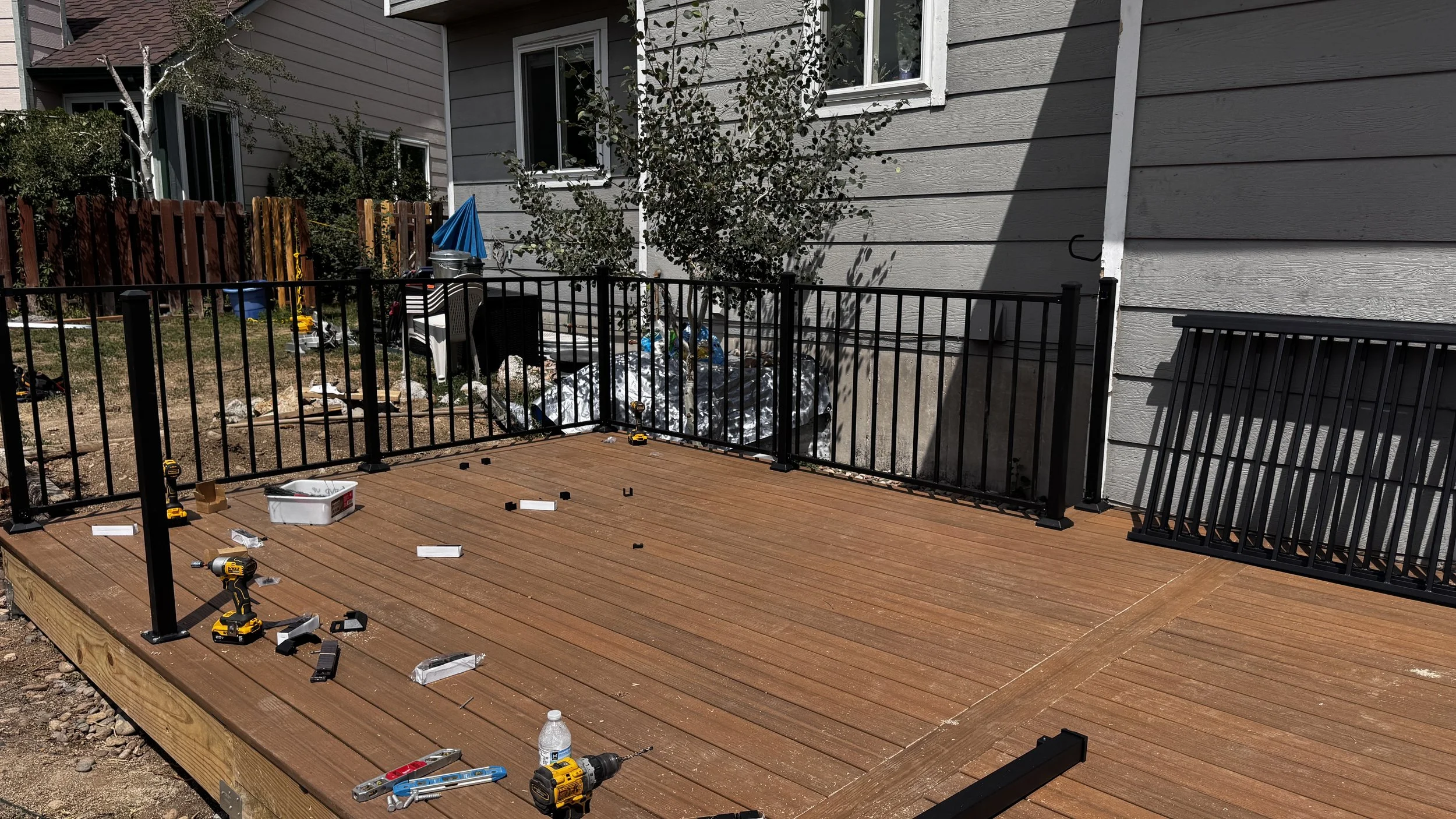 Construction of a wooden deck with a black metal safety fence being assembled. Tools, screws, and materials are scattered on the deck. The background shows a house with gray siding, a window, and a small tree.