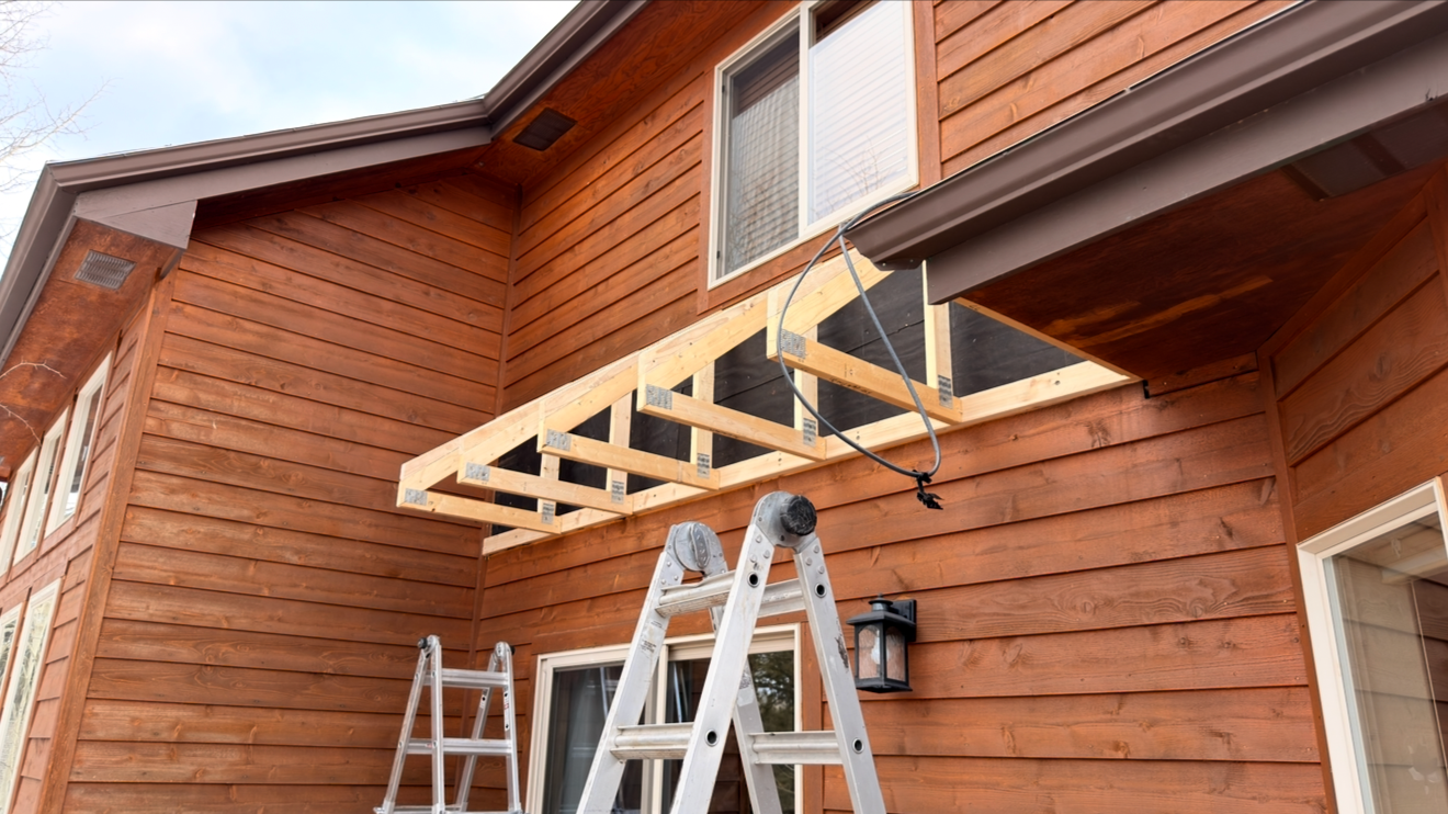 Front of a wooden house under construction with a partially built balcony, ladders, and construction tools
