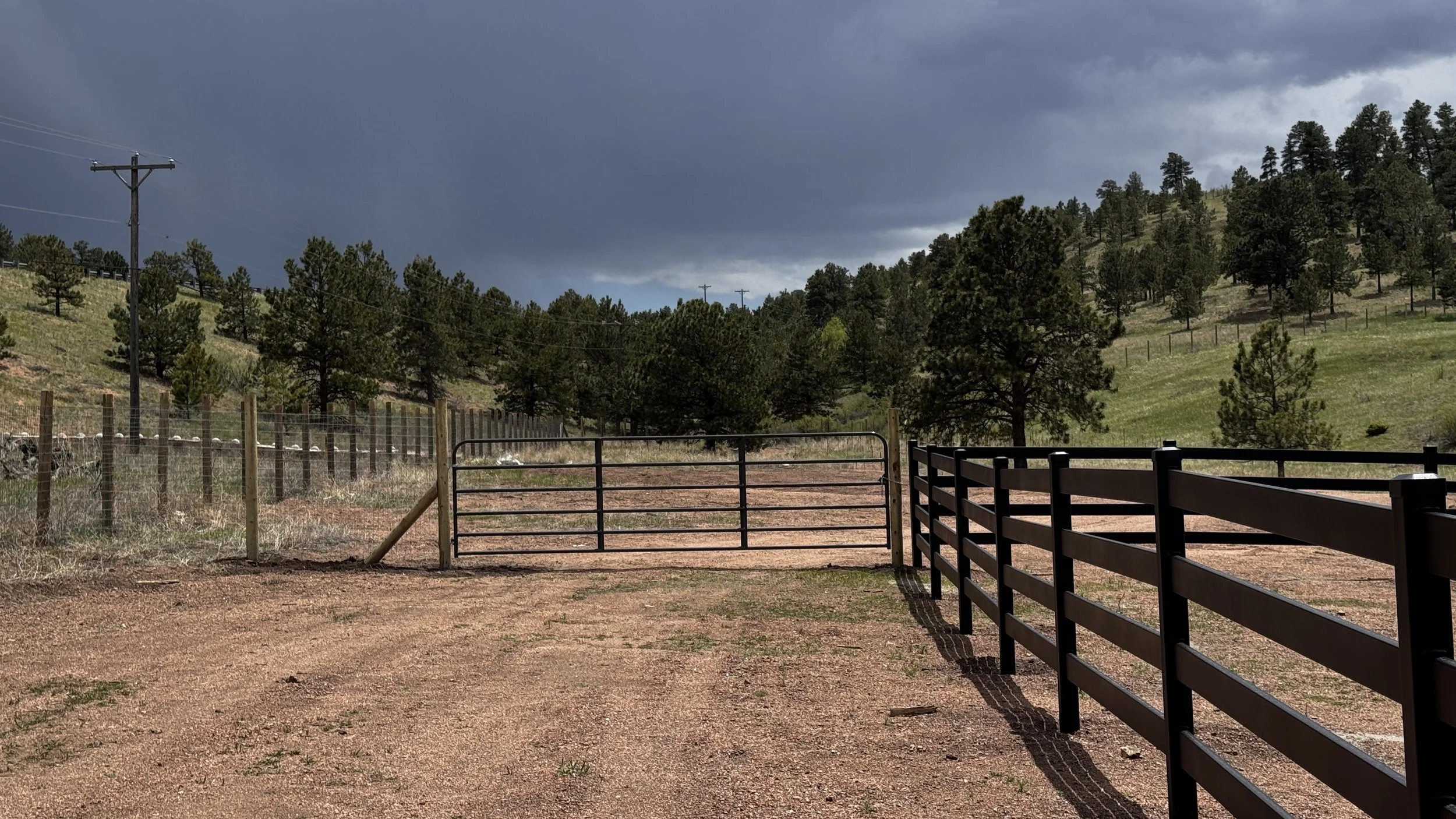 View of a fenced area with black metal gates, dirt ground, green trees on rolling hills, and a dark, cloudy sky in the background.