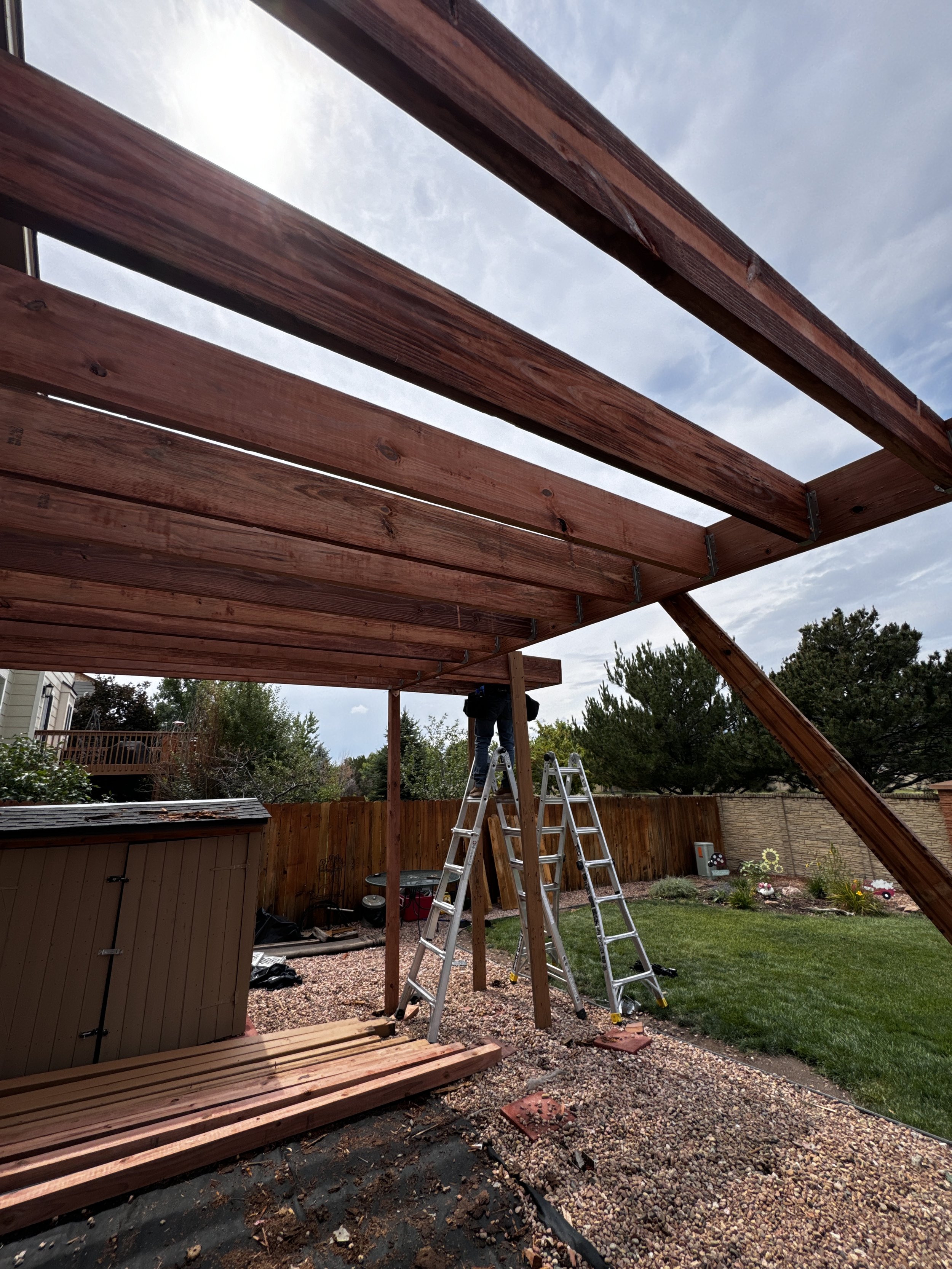Construction of a wooden deck in a backyard, with ladders and materials on the ground, and partially built wooden beams overhead.