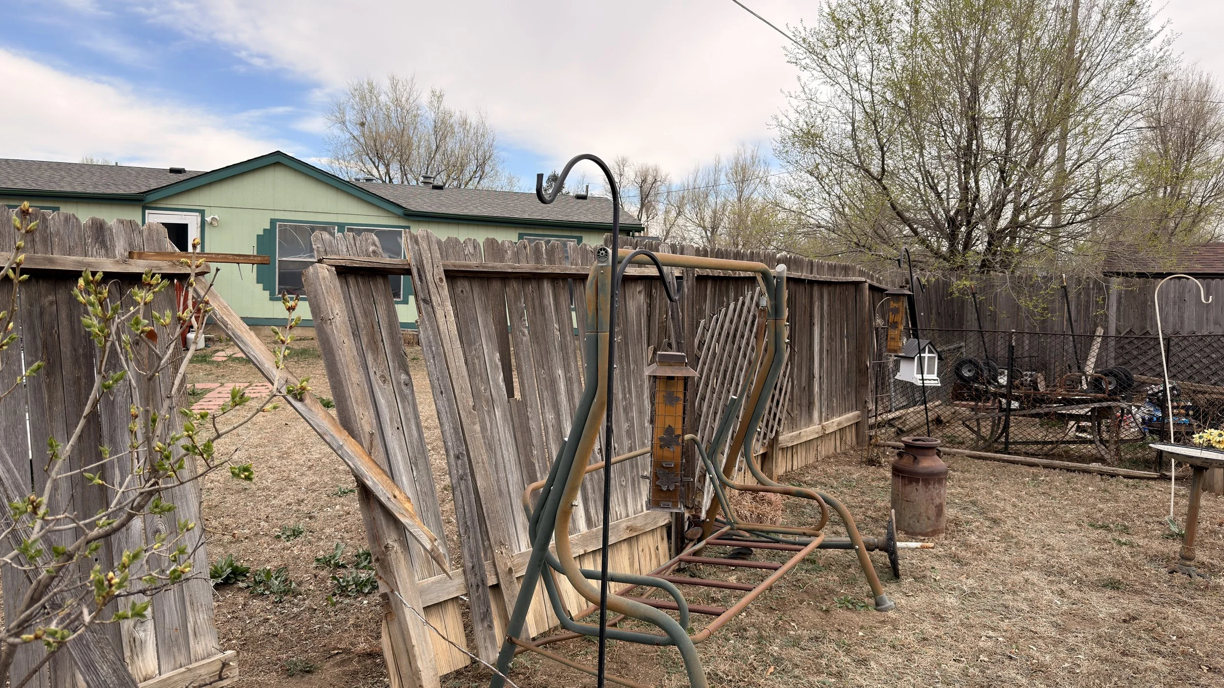 A backyard with a dilapidated wooden fence, old garden equipment, a leafless tree, and a green house with a gray roof in the background under a partly cloudy sky. Repair post