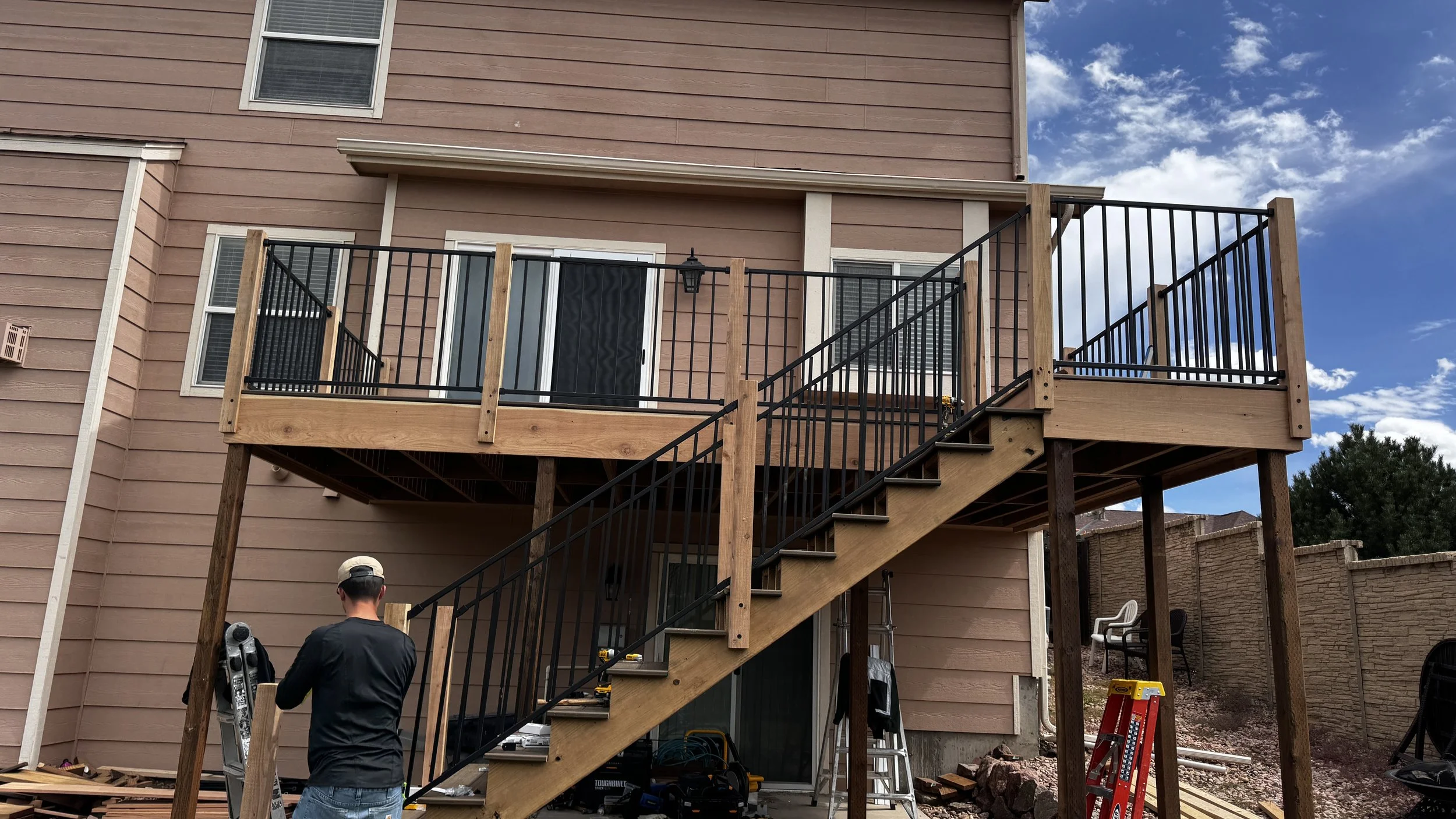 A backyard with a newly constructed wooden deck attached to the back of a two-story house with pink siding. The deck has black metal railings and stairs leading down to the yard, where two workers are present. The sky is partly cloudy.
