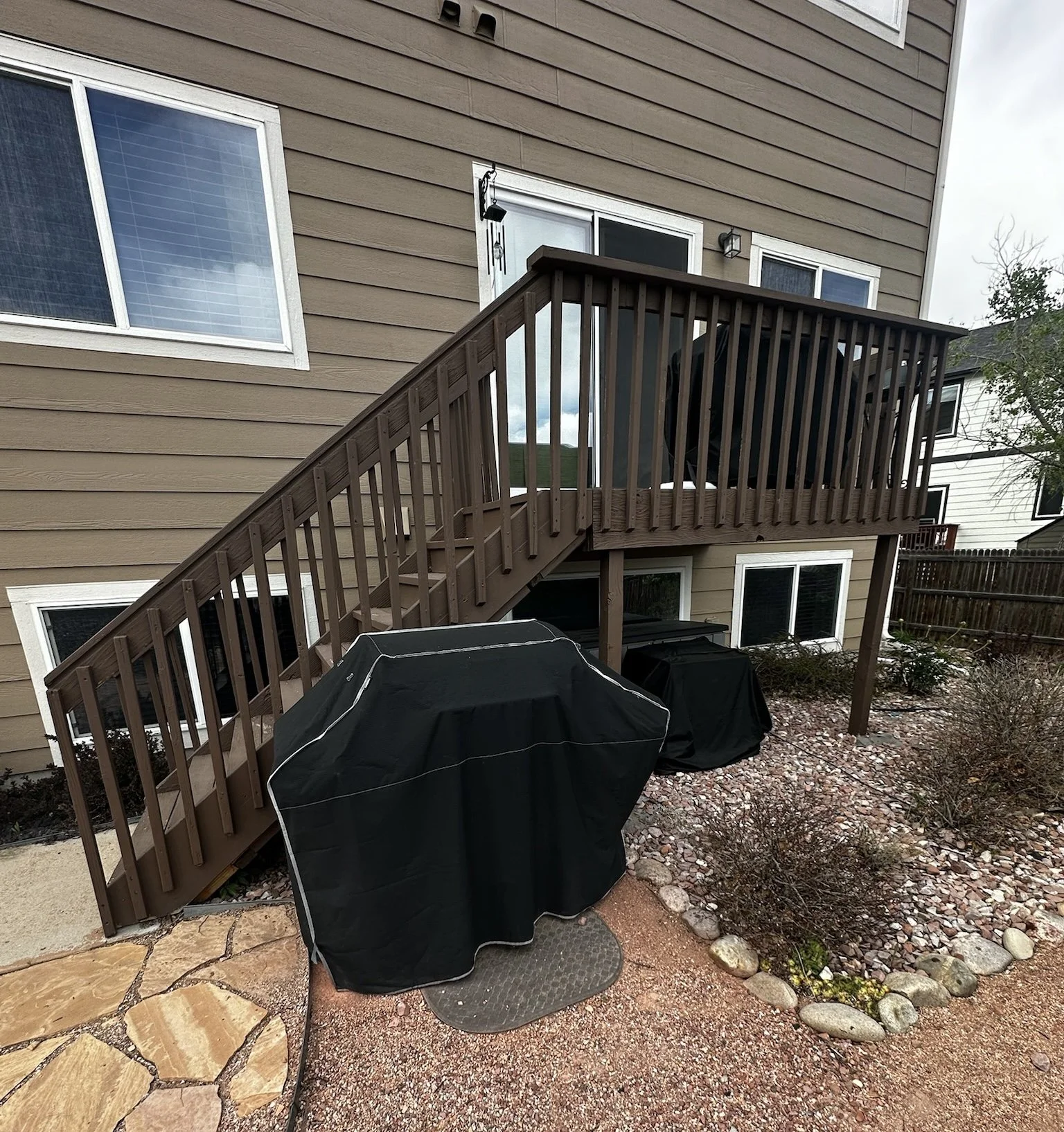 Backyard with a house featuring beige siding, a small wooden deck with a staircase, two closed grills with covers, a patio area with stone pavers, and a landscaped garden with rocks and bushes.